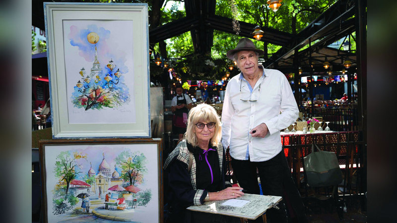 Polish watercolourist artist Marie Jolann «Yola» and her french husband Francois Renouf de Boyrie pose for photos next to her art works at Place du Tertre, the famous painters' square on the hill in the Montmartre district, northern Paris, on July 17, 2024. On the Place du Tertre, artists but there is no rush of tourists and the restaurant terraces are rather empty, just a few days before the 2024 Paris Olympics Games. (Photo by Miguel MEDINA / AFP)